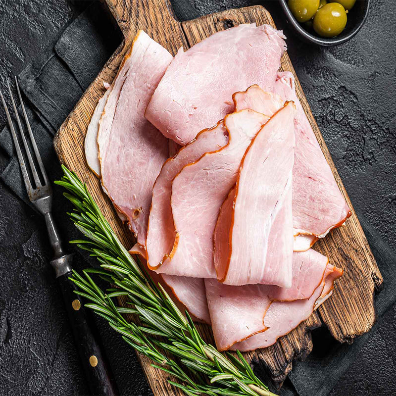 Sliced ham on a wooden board with rosemary and olives on a dark surface